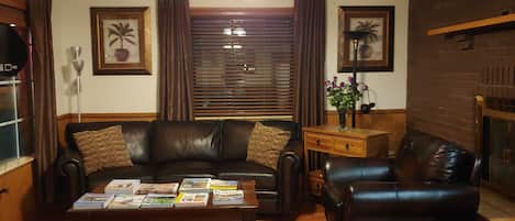 A living room with a brown leather sofa, a coffee table with magazines, a wooden side table, and a ceiling fan.