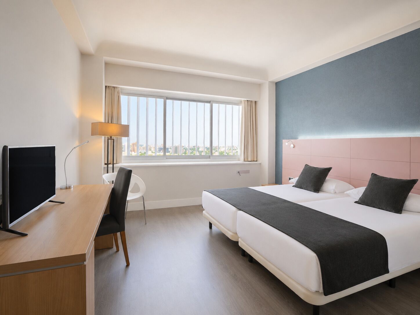 Habitación de hotel con cama, escritorio con computadora, silla, ventana con cortinas y vista a la ciudad.