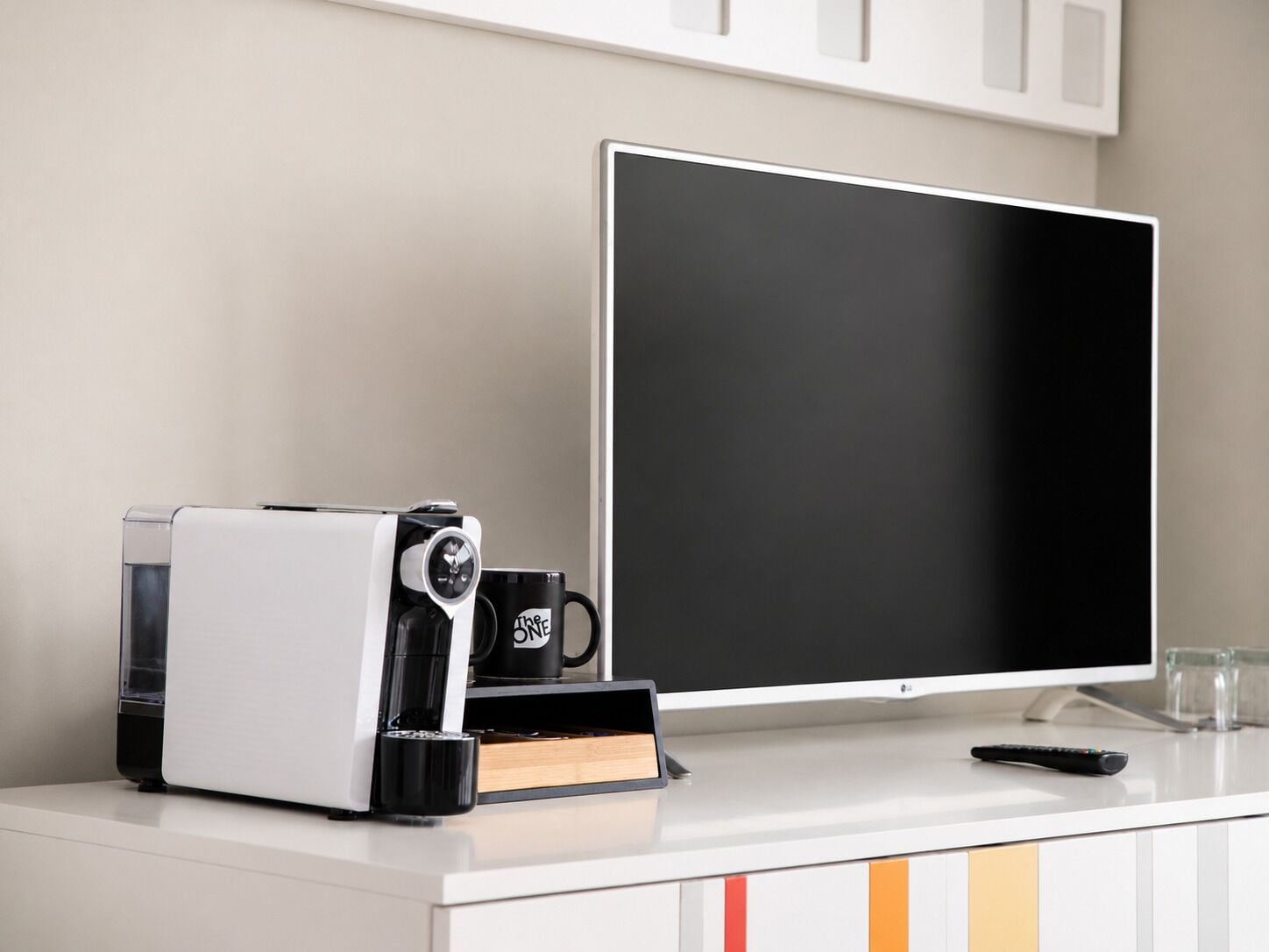 A coffee machine, a mug, a remote control, and a computer monitor on a desk.