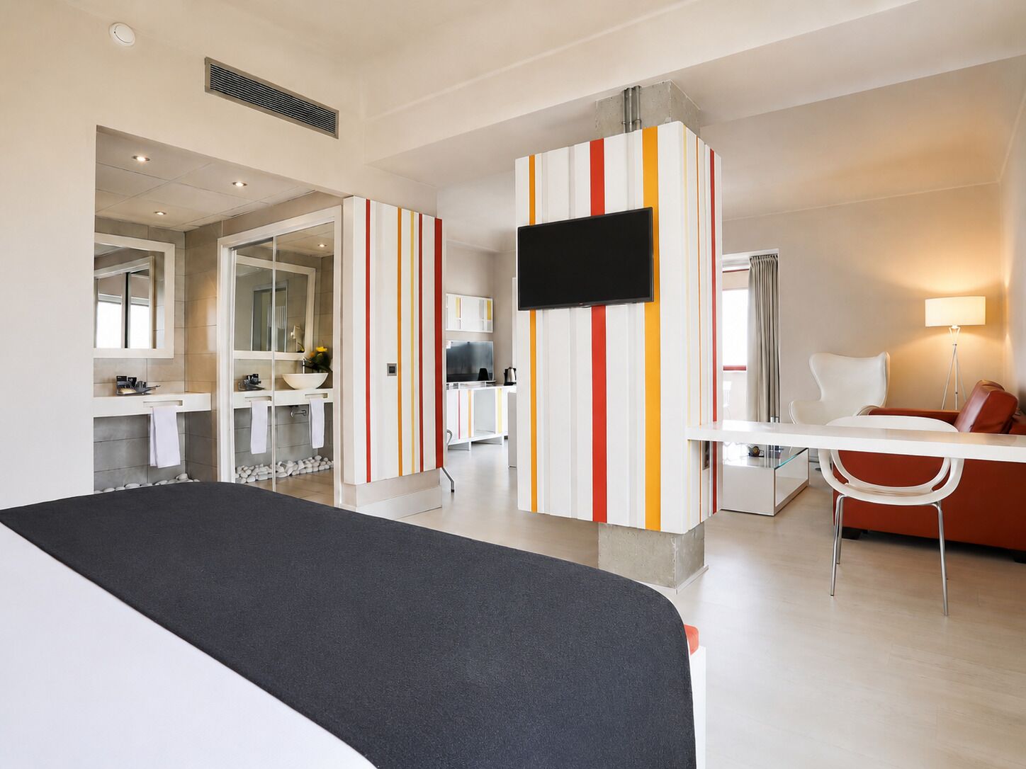 A modern hotel room with a bed, a TV, a desk, a chair, and a bathroom with a mirror and sink.