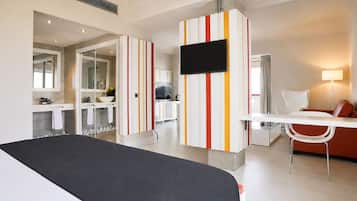 A modern hotel room with a bed, a TV, a desk, a chair, and a bathroom with a mirror and sink.