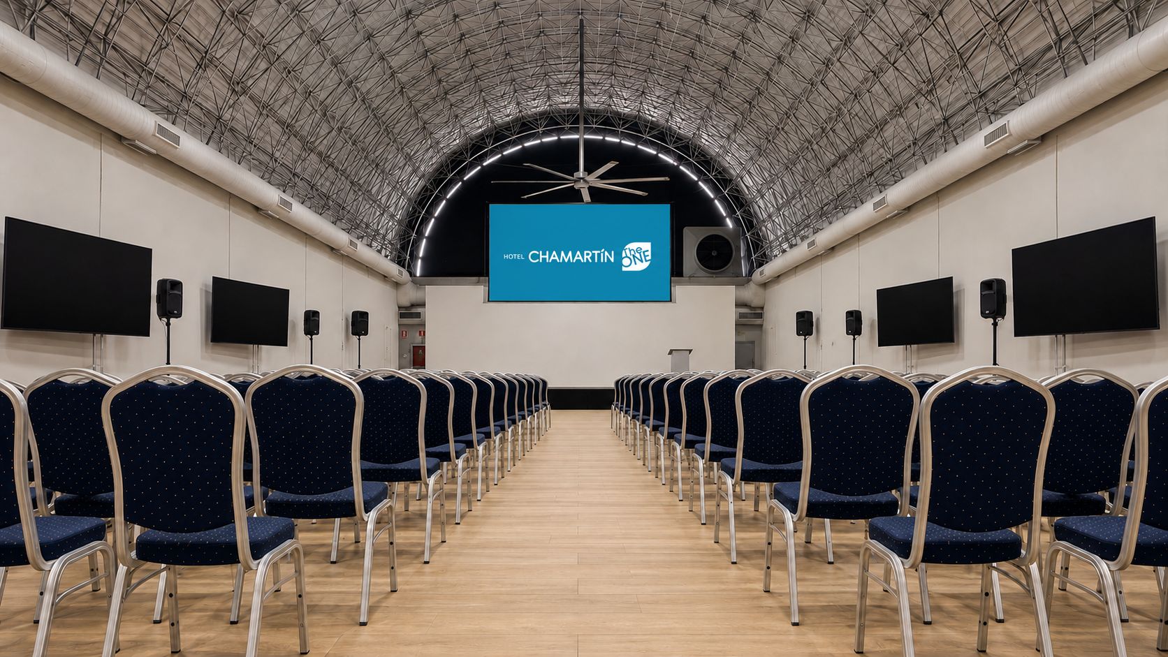 Een congreszaal met rijen stoelen, een groot scherm aan de voorkant en een hoog plafond met een metalen constructie.