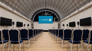 A conference room with rows of chairs, a large screen at the front, and a high ceiling with a metal framework. - Hotel Chamartin The One (Madrid)