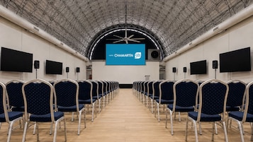 A conference room with rows of chairs, a large screen at the front, and a high ceiling with a metal framework.