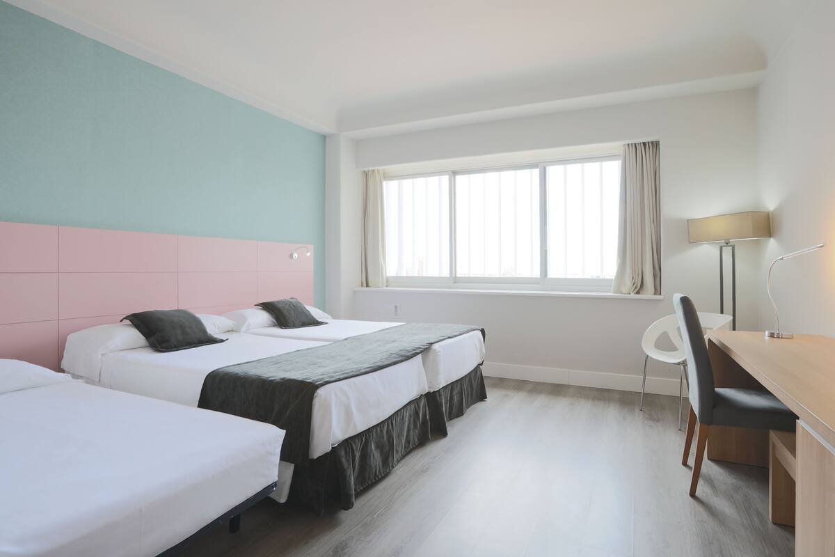 A hotel room with two beds, a desk, and a chair. There is a window with curtains, and the room has a light-colored floor.