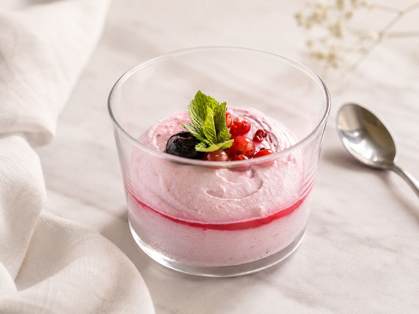 A clear glass dessert bowl with a layered dessert, garnished with berries and mint leaves, on a white surface with a spoon beside it.