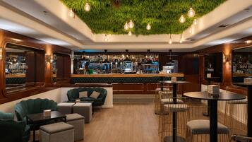 A bar area with a green wall, wooden bar counter, and seating with round tables and chairs.