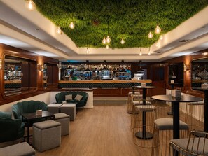 Zona de bar con pared verde, mostrador de madera y asientos con mesas redondas y sillas.
