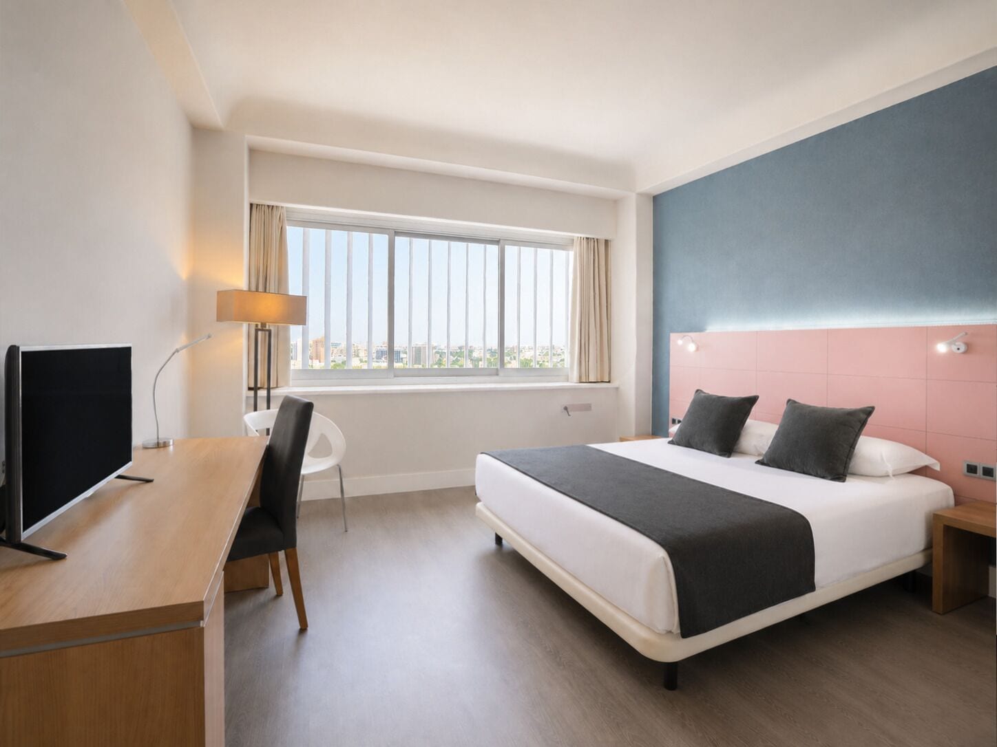 Habitación de hotel con cama, escritorio, silla, televisión y ventana con cortinas.