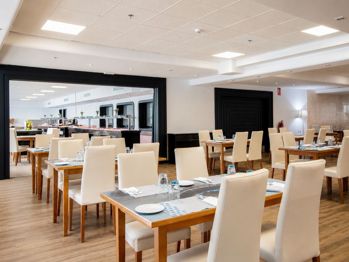 Een moderne eetruimte in een hotel met meerdere tafels en stoelen, een zichtbare keuken en een grote zwarte deur.