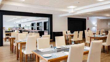 Een moderne eetruimte in een hotel met meerdere tafels en stoelen, een zichtbare keuken en een grote zwarte deur.