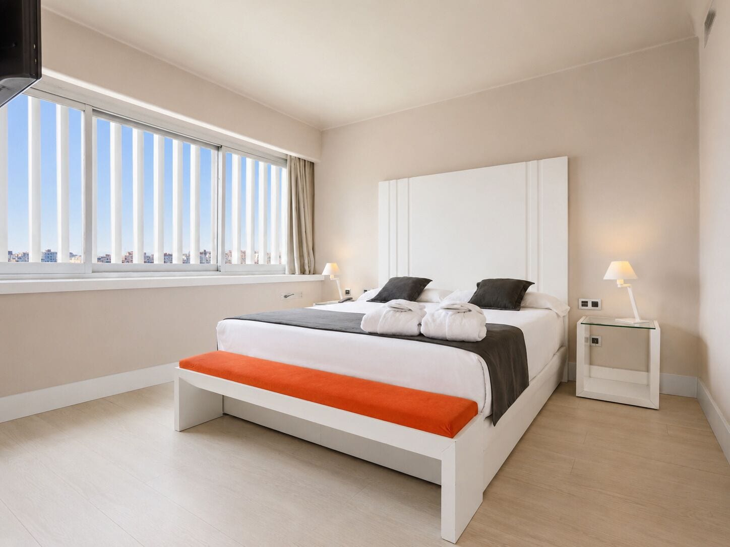 A modern hotel room with a large bed, two bedside tables, a window with blinds, and a city view.