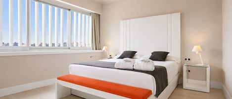 A modern hotel room with a large bed, two bedside tables, a window with blinds, and a city view.