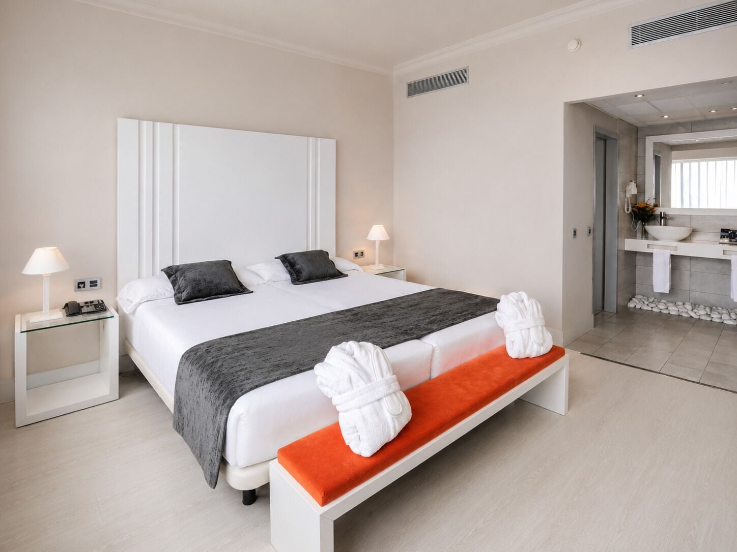 Habitación de hotel con una cama grande, mesitas de noche, un banco y un baño visible a través de una puerta abierta.
