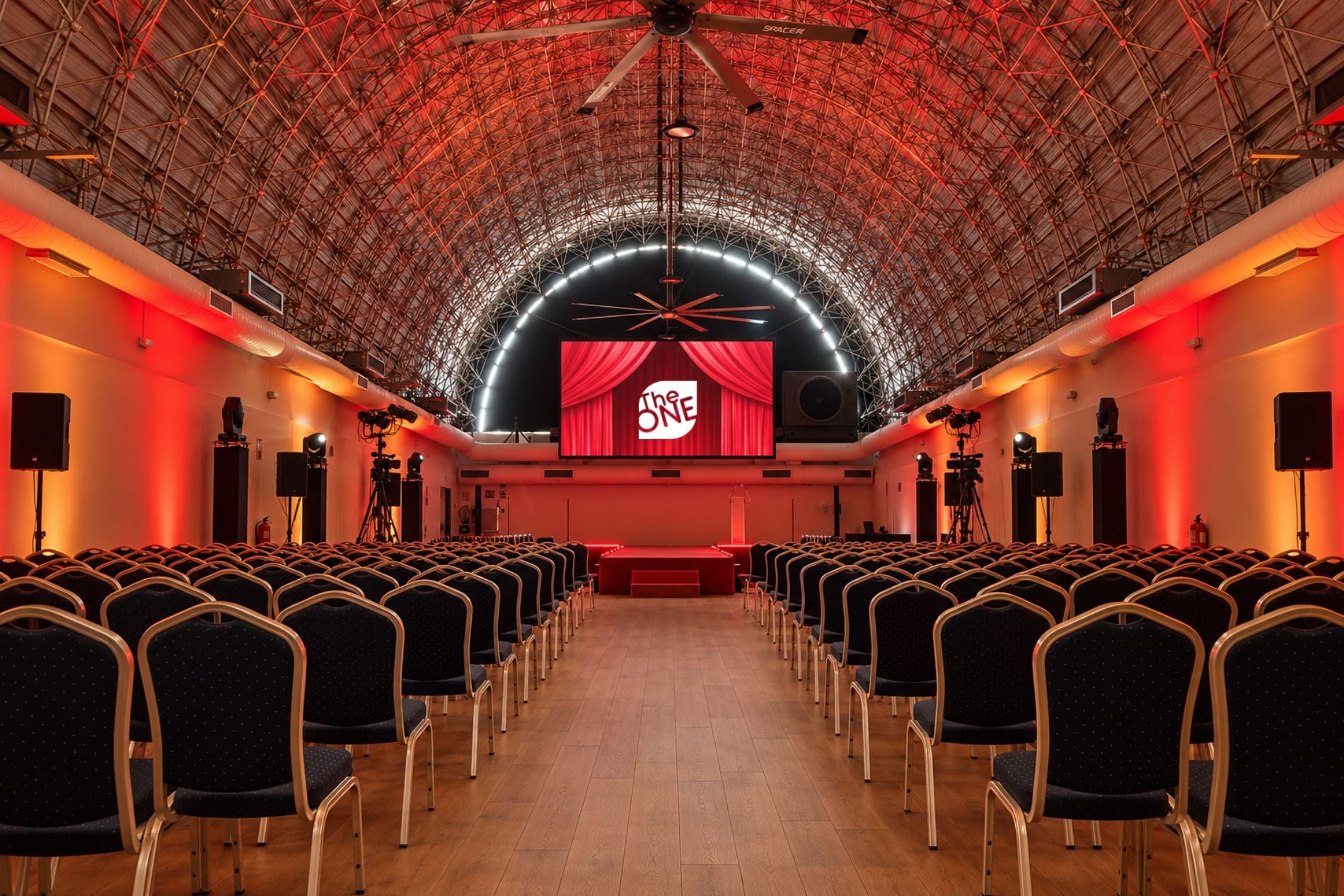 Un auditorio con filas de sillas, un escenario con cortina roja y una pantalla grande mostrando 'The ONE'.
