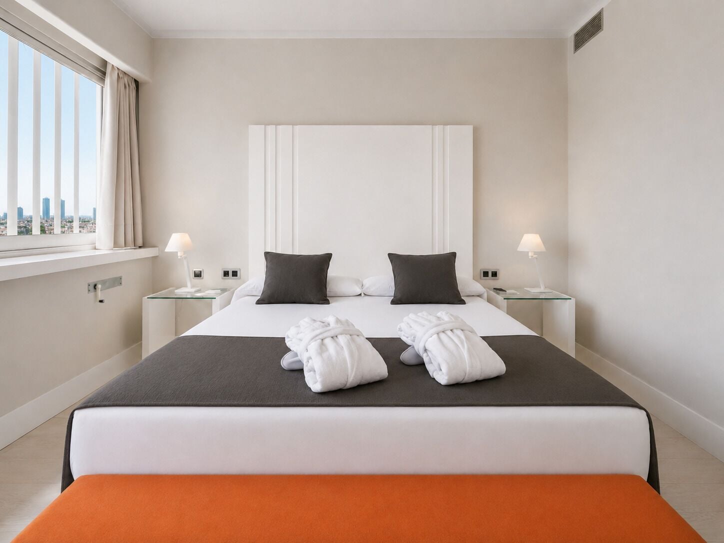 Una camera d'albergo con un letto grande, due comodini con lampade, una finestra con tende e vista sulla città.