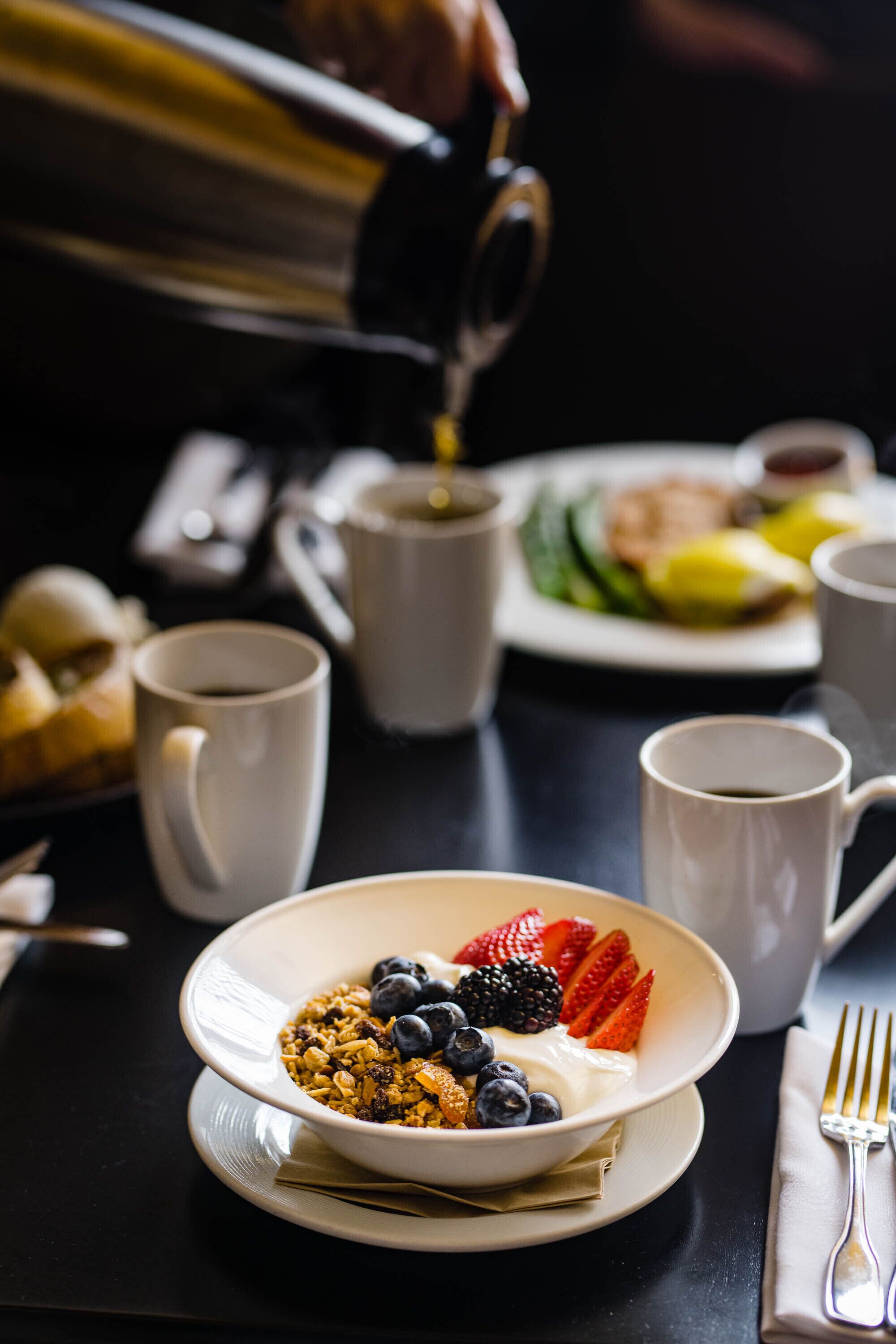 Une table mise pour le petit-déjeuner, avec un bol de céréales garni de fruits, une tasse de café et une assiette de nourriture en arrière-plan.