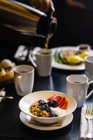 A breakfast setting with a bowl of cereal topped with fruits, a coffee cup, and a plate of food in the background.
