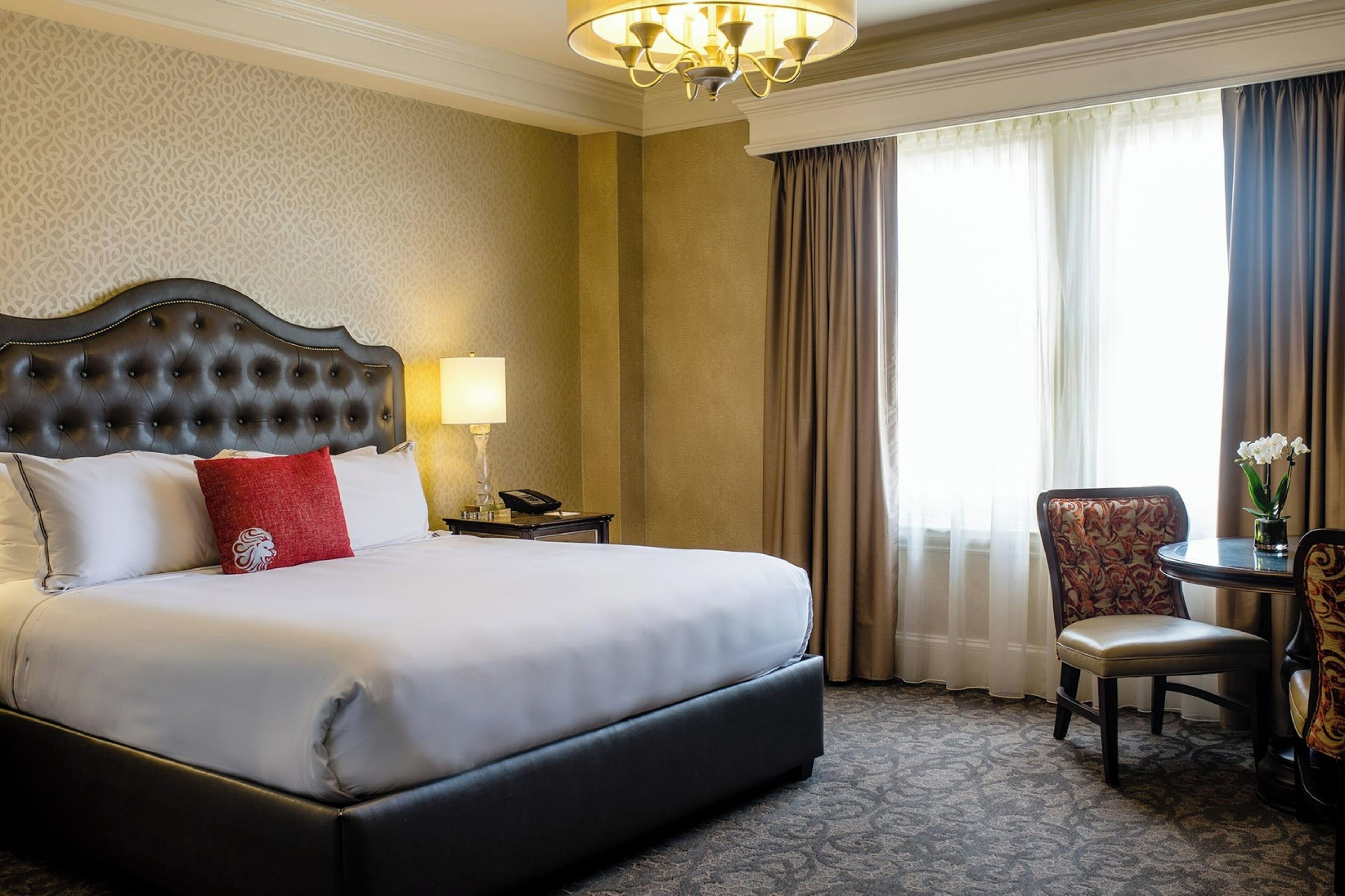 A hotel room with a large bed, a bedside table with a lamp, a chair, and a view of the outside through curtains.