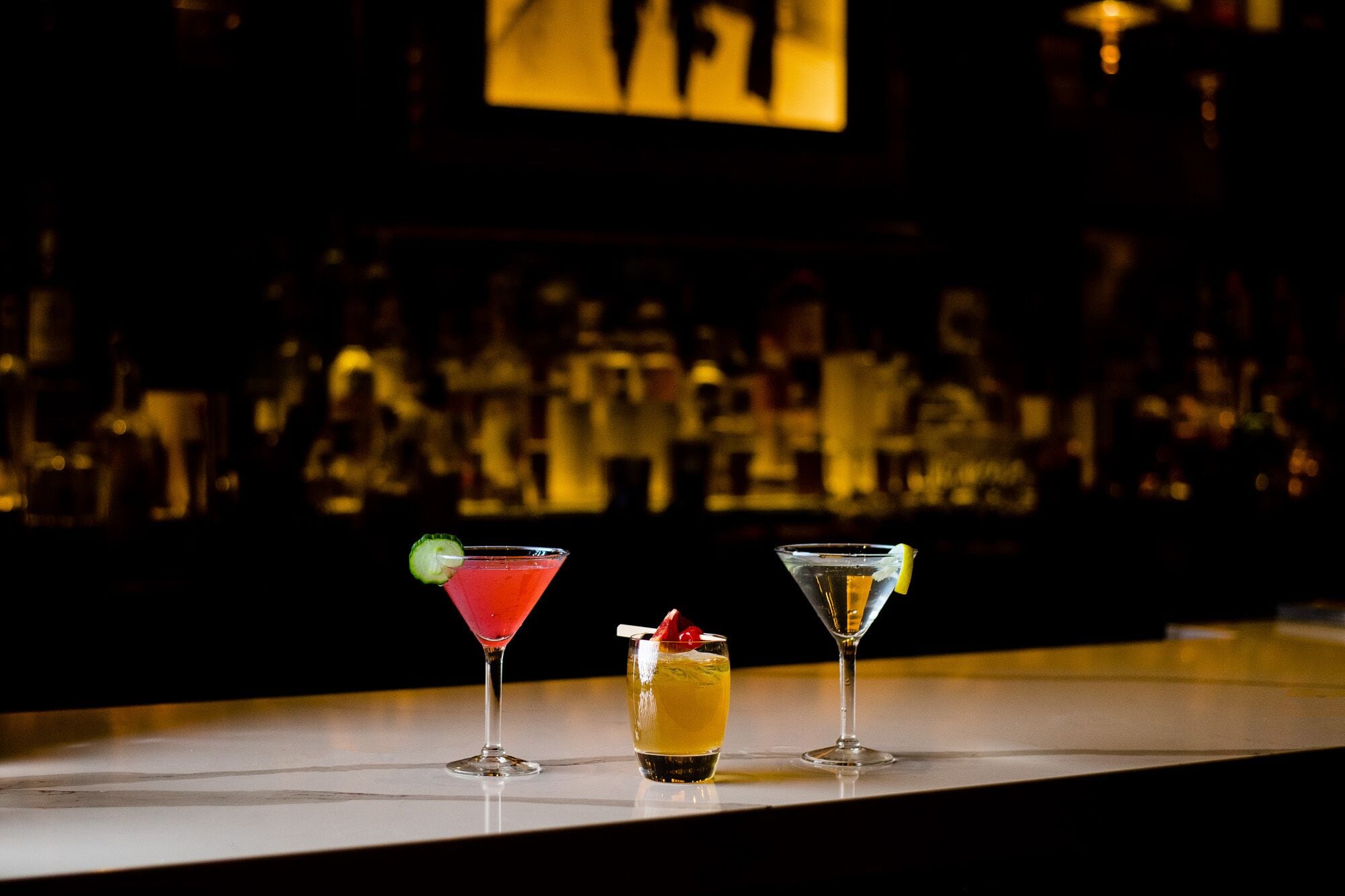 Drei Cocktails auf einer Bar, im Hintergrund verschwommene Flaschen.