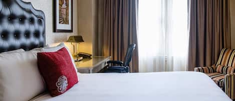 Habitación de hotel con una cama grande, un escritorio con lámpara, una silla y un ventanal con cortinas.