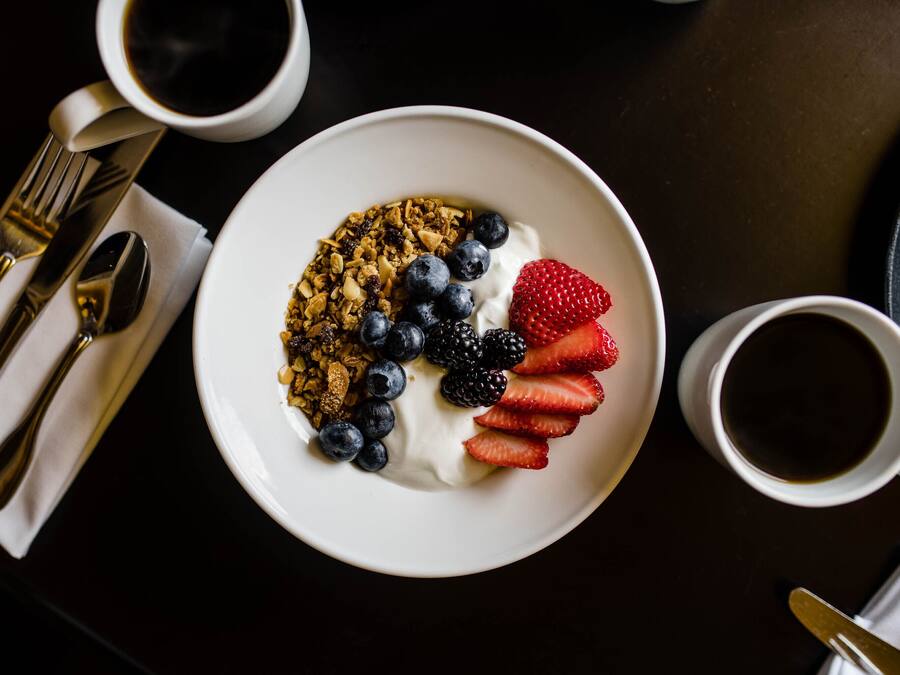 Un plato blanco con yogur, granola, arándanos, zarzamoras y fresas sobre una mesa oscura.