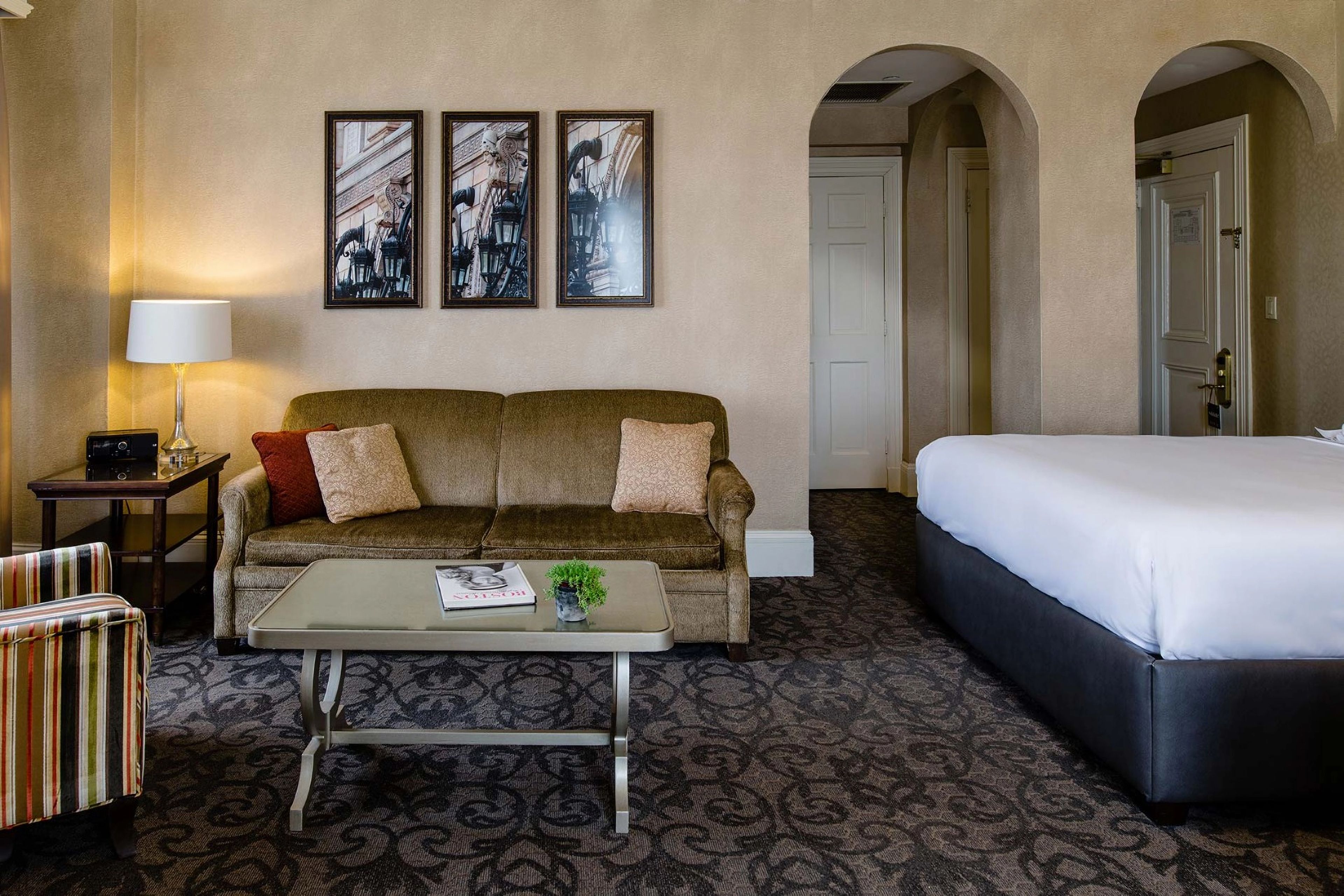 Une chambre d’hôtel avec un canapé, un lit, une table basse, une lampe et des tableaux encadrés au mur.