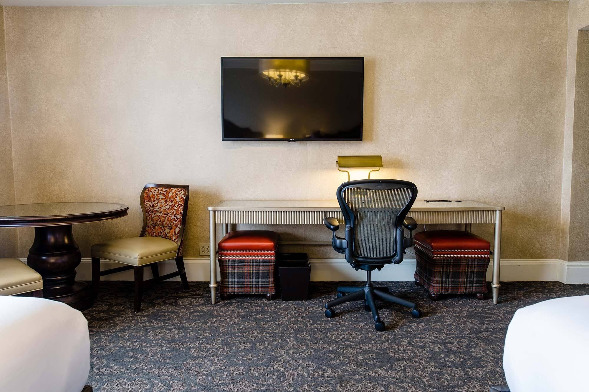 Ein Hotelzimmer mit Flachbildfernseher an der Wand, einem kleinen Schreibtisch mit Lampe, zwei karierten Hocker, einem gemusterten Sessel und einem runden Tisch mit Stuhl.