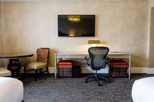 Ein Hotelzimmer mit Flachbildfernseher an der Wand, einem kleinen Schreibtisch mit Lampe, zwei karierten Hocker, einem gemusterten Sessel und einem runden Tisch mit Stuhl.