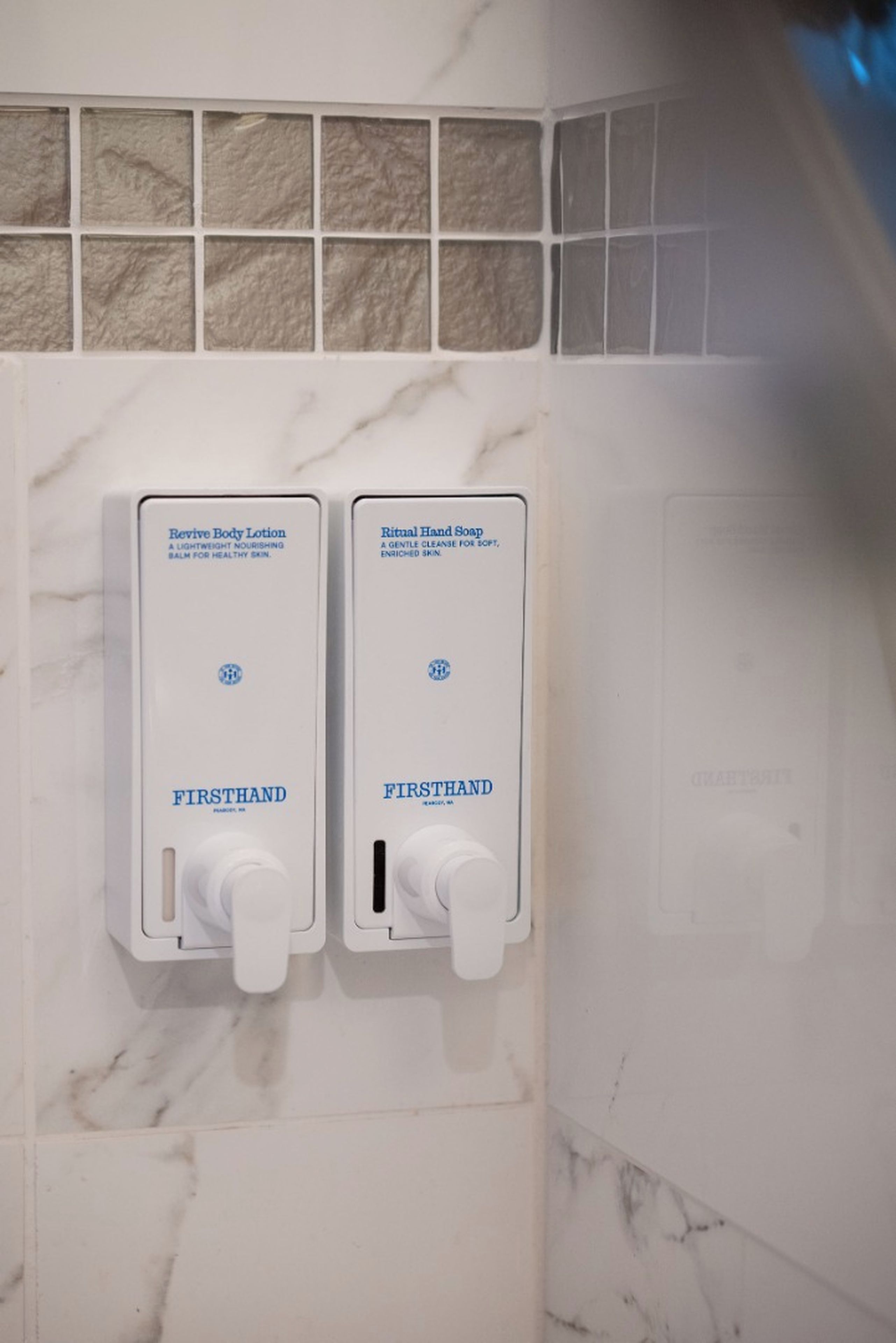 A bathroom with a marble wall and two dispensers labeled 'Revive Body Lotion' and 'Ritual Hand Soap' by Firsthand.