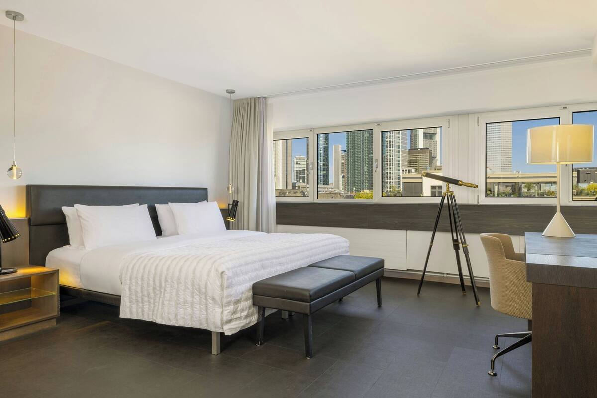 A modern hotel room with a large bed, a desk, and a view of the cityscape.