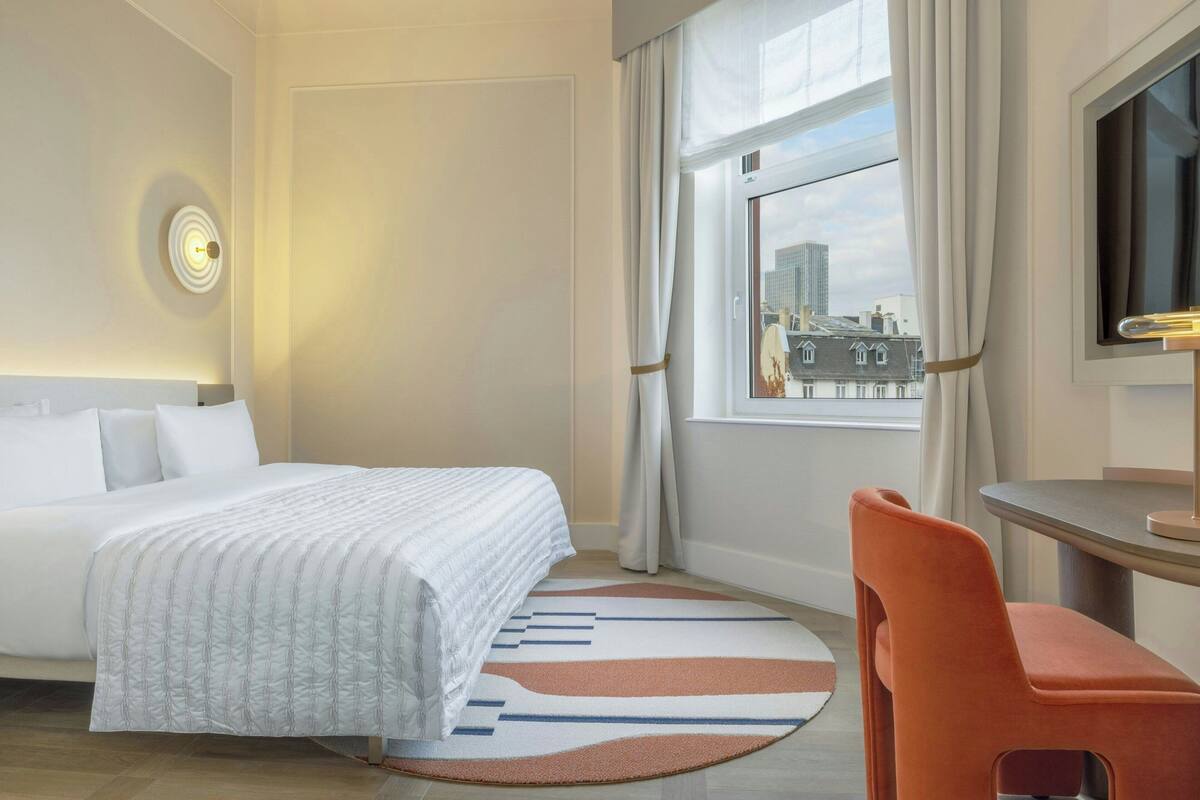 A hotel room with a bed, a desk, a chair, a large window with curtains, and a city view.