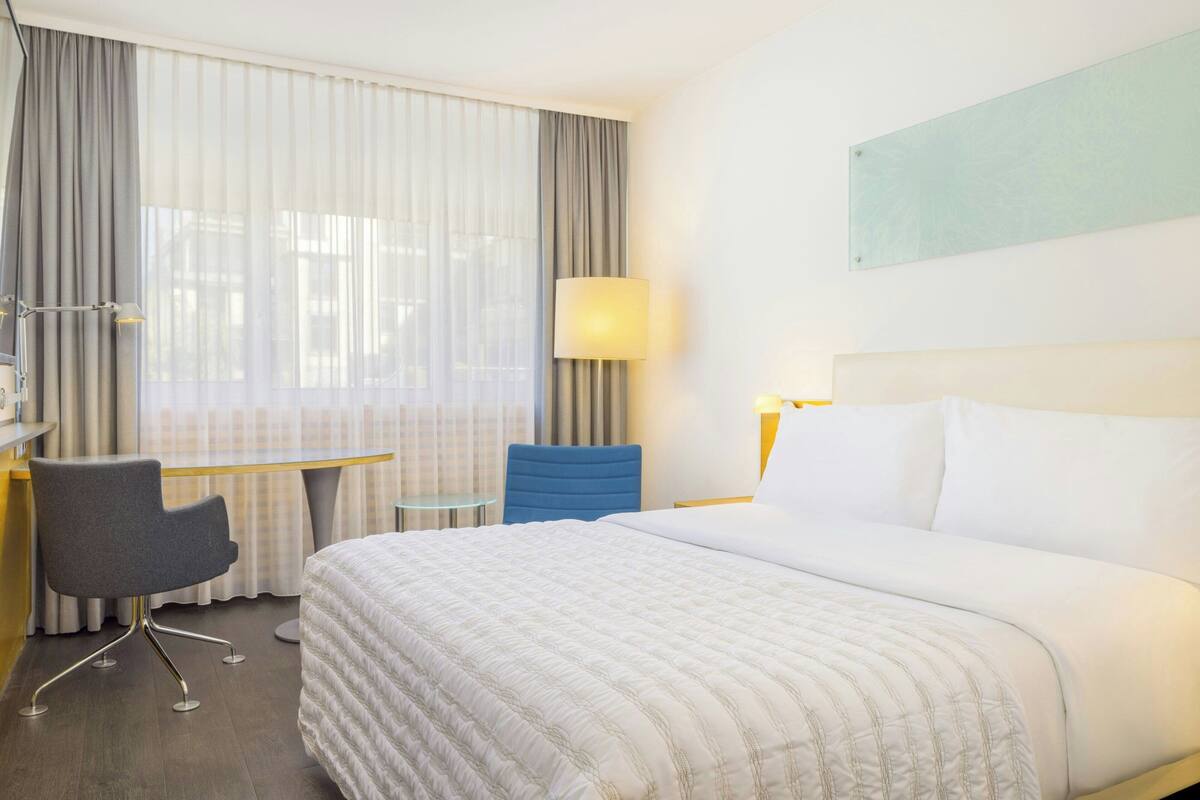 A hotel room with a large bed, a desk with a chair, a lamp, and a view through sheer curtains.