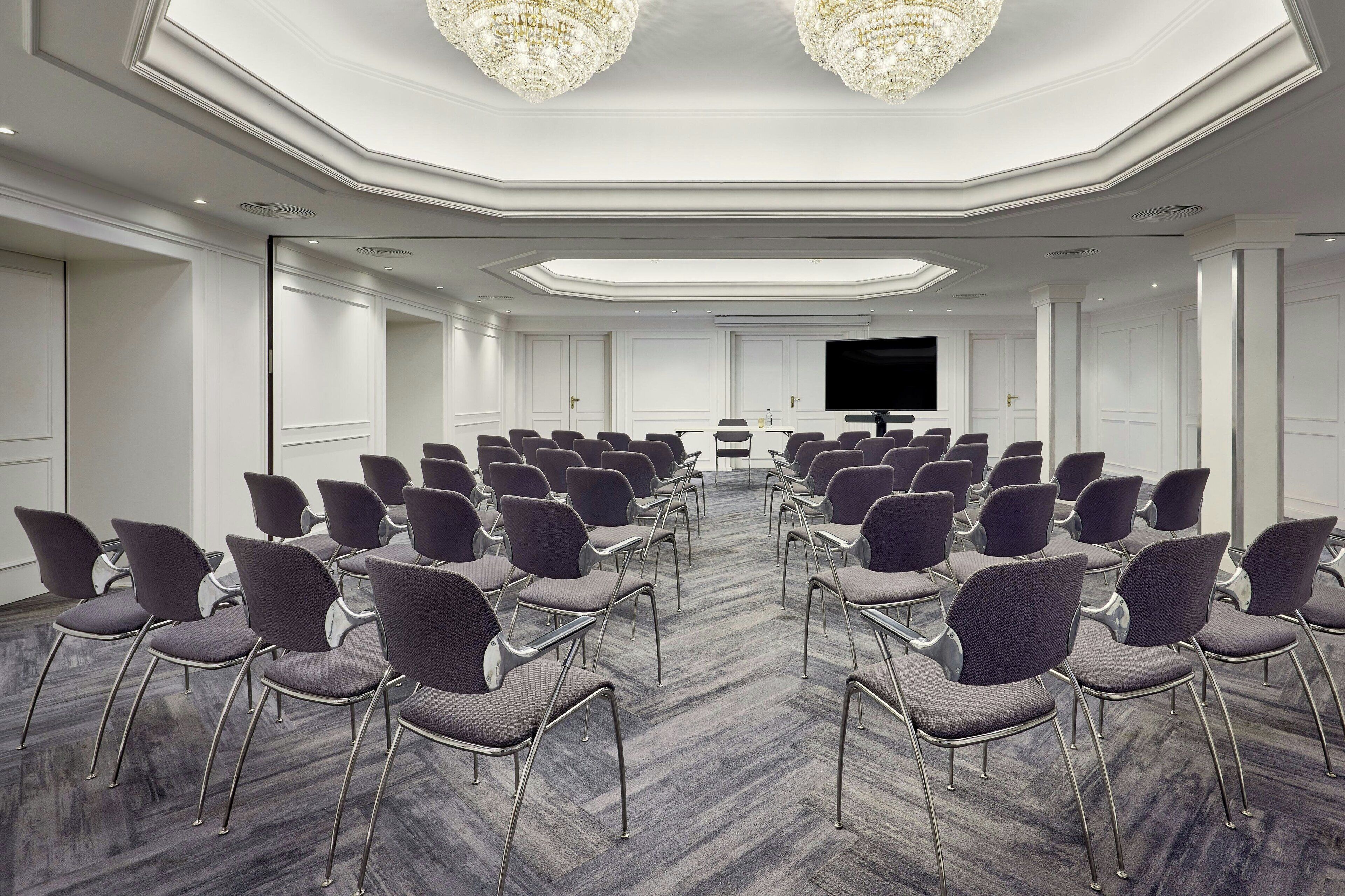 A conference room with rows of chairs facing a large screen, a chandelier, and a modern ceiling design.