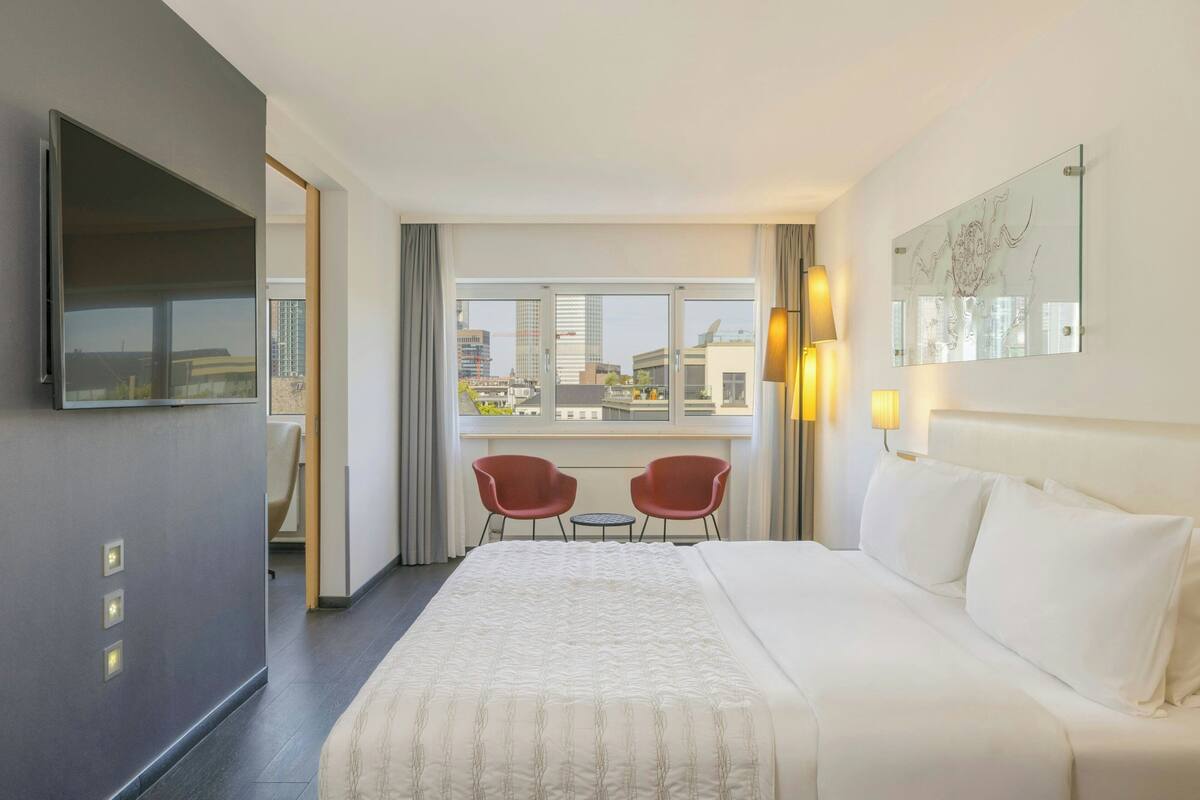A hotel room with a large bed, a TV, two red chairs, a window with a city view, and a wall-mounted artwork.