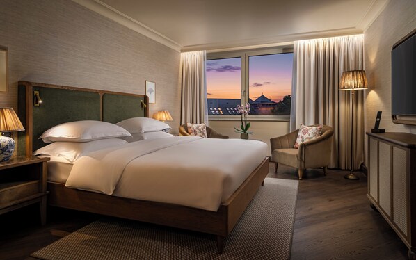 A hotel room with a large bed, a sitting area with a chair, a desk, and a TV.