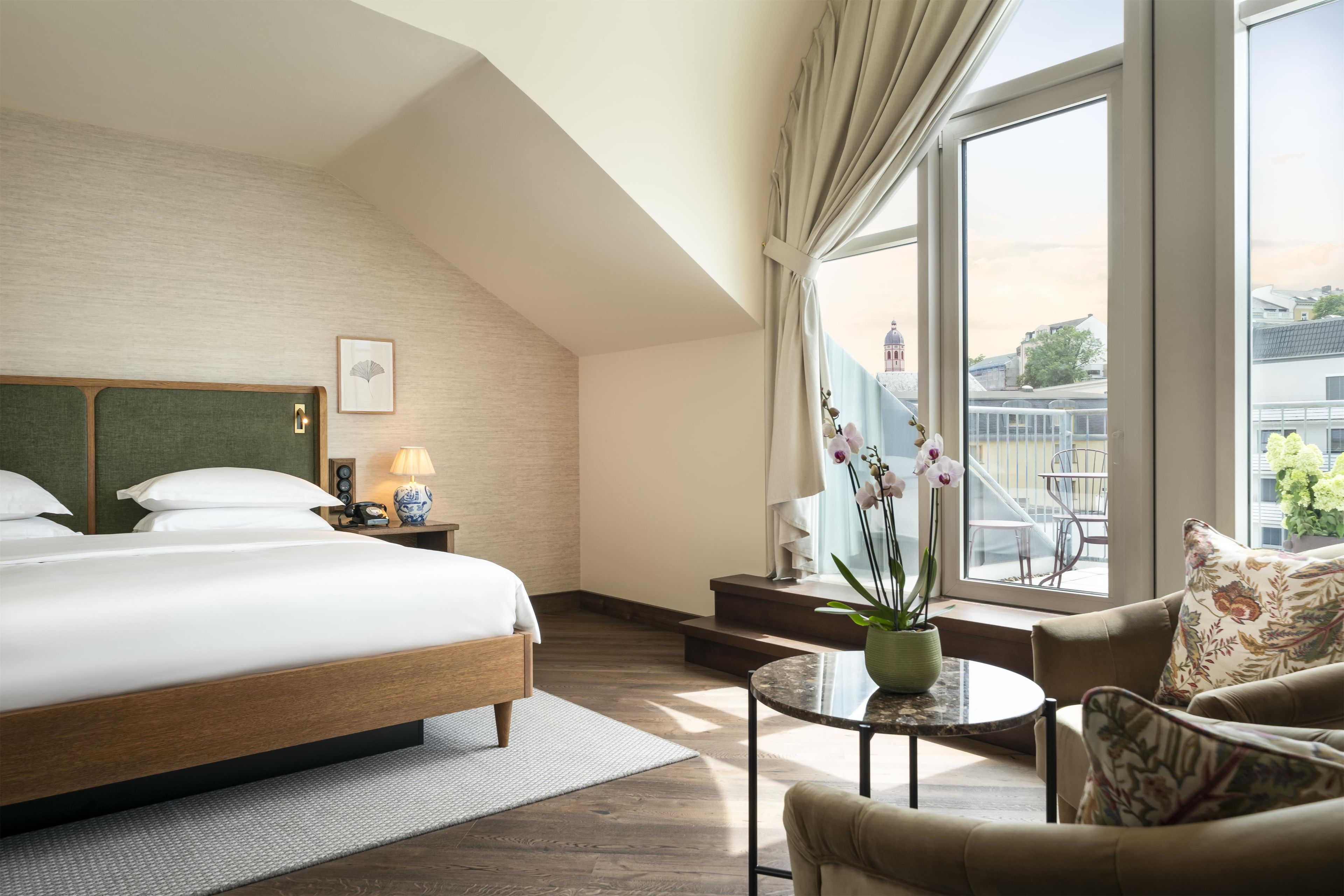 Una camera d'albergo con un letto grande, un angolo salotto con tavolo e sedie, vista sulla città dalla finestra e una scala che conduce a un balcone esterno.