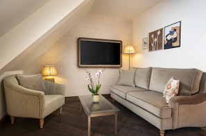 Una sala de estar acogedora con un sofá, un sillón y una mesa de centro. Un televisor de pantalla plana está montado en la pared, y hay dos lámparas de pared.