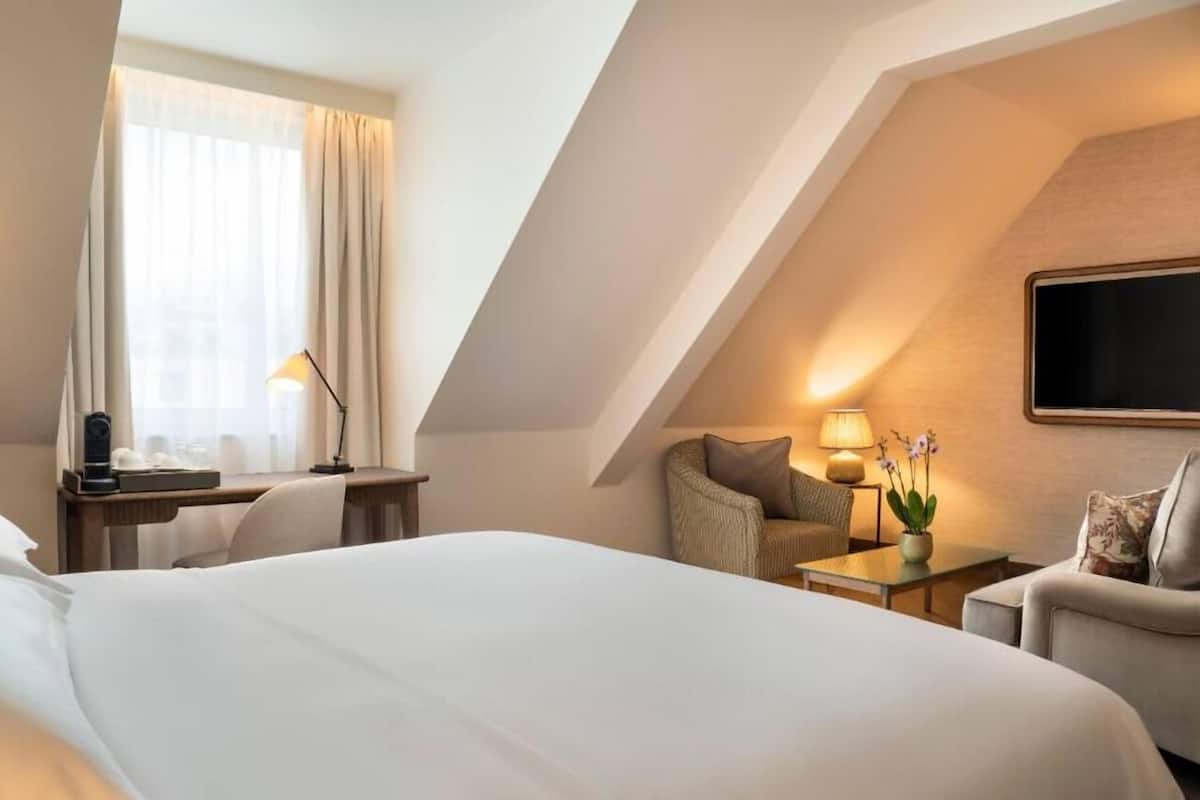 Habitación de hotel con una cama grande, un escritorio con lámpara, una silla, un sofá y un televisor montado en la pared.