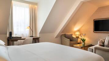 A hotel room with a large bed, a desk with a lamp, a chair, a sofa, and a TV mounted on the wall.