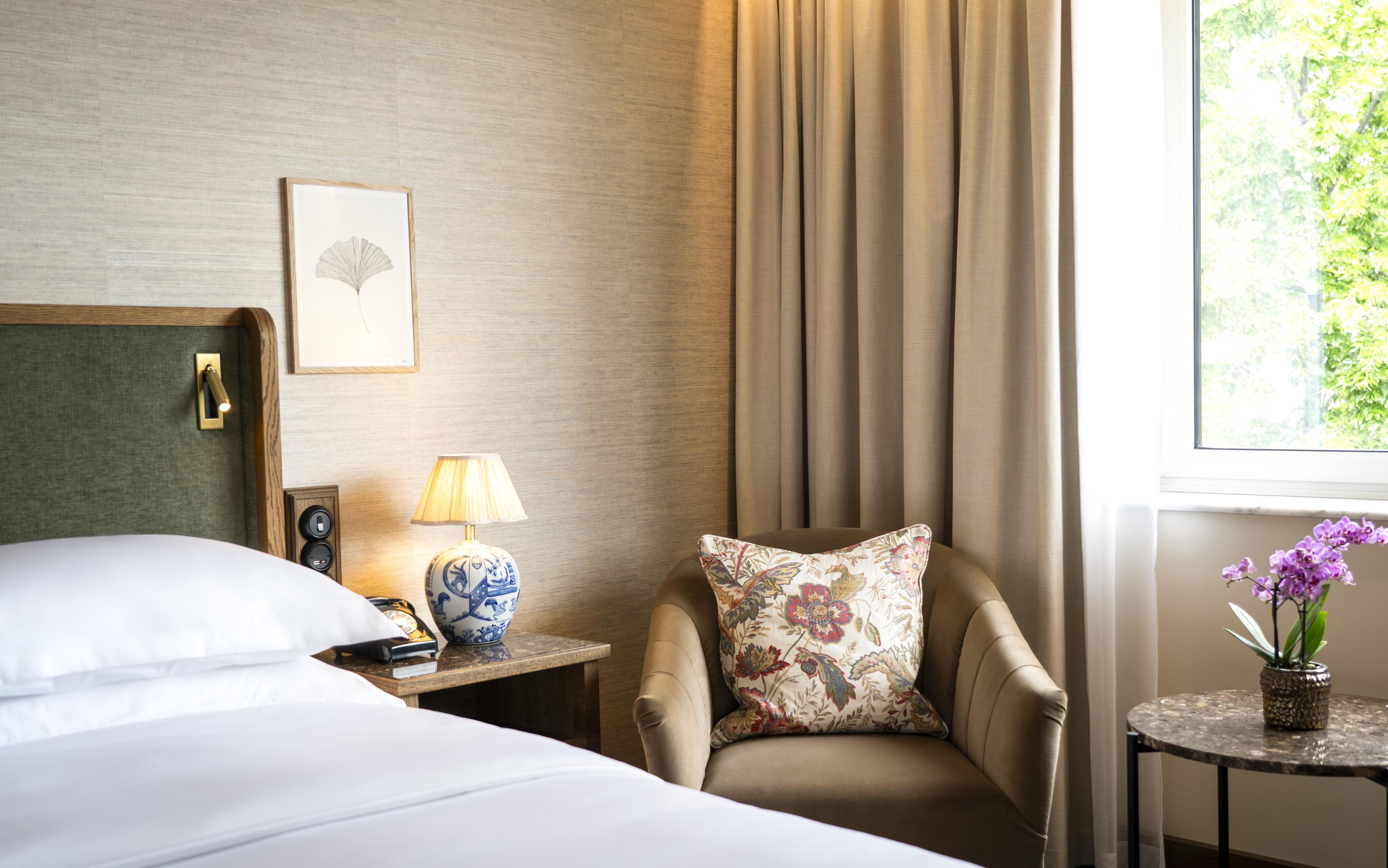 Een hotelkamer met een bed, een stoel, een nachtkastje met lamp, een lijst met foto aan de muur en een raam met gordijnen.