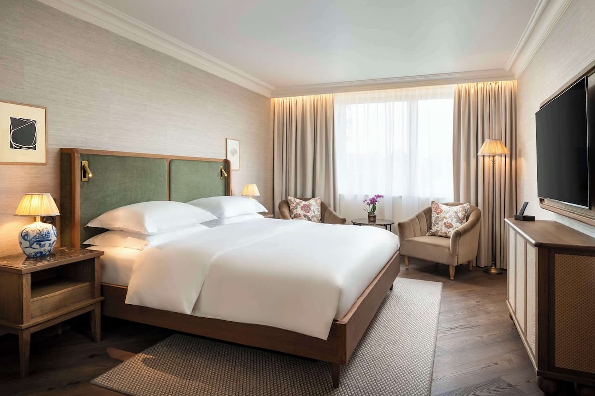 Una habitación de hotel con una cama grande, televisión, escritorio con lámpara y una silla con cojín floral.