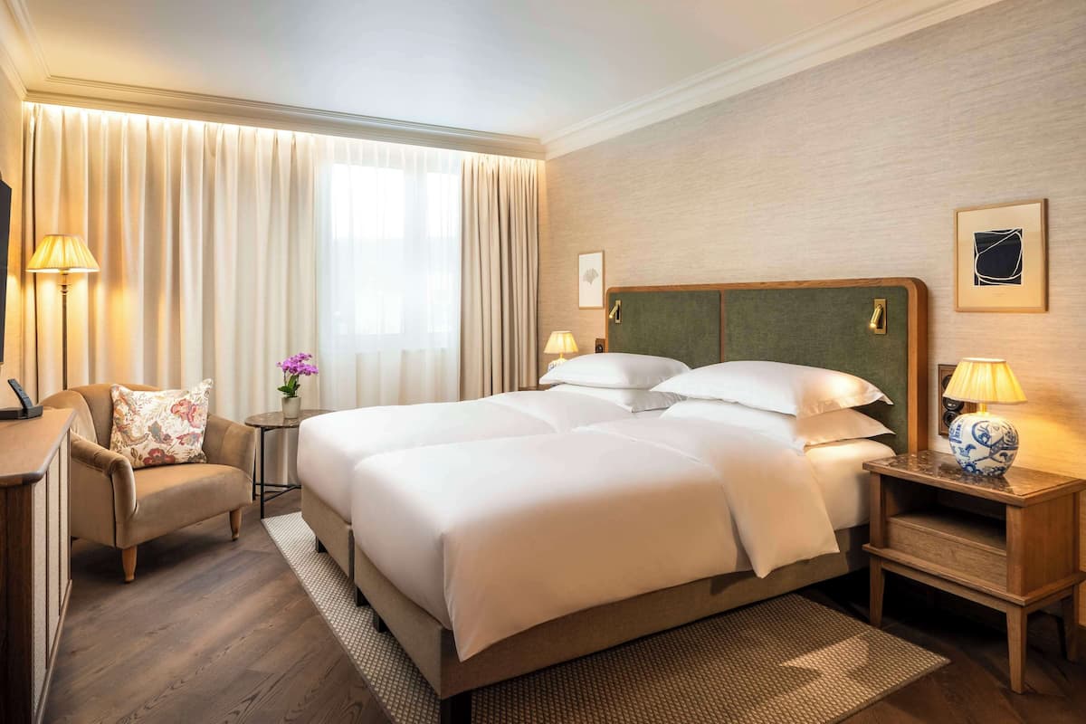 Habitación de hotel con una cama grande, dos mesitas de noche con lámparas, una silla, un escritorio con televisor y un ventanal con cortinas.