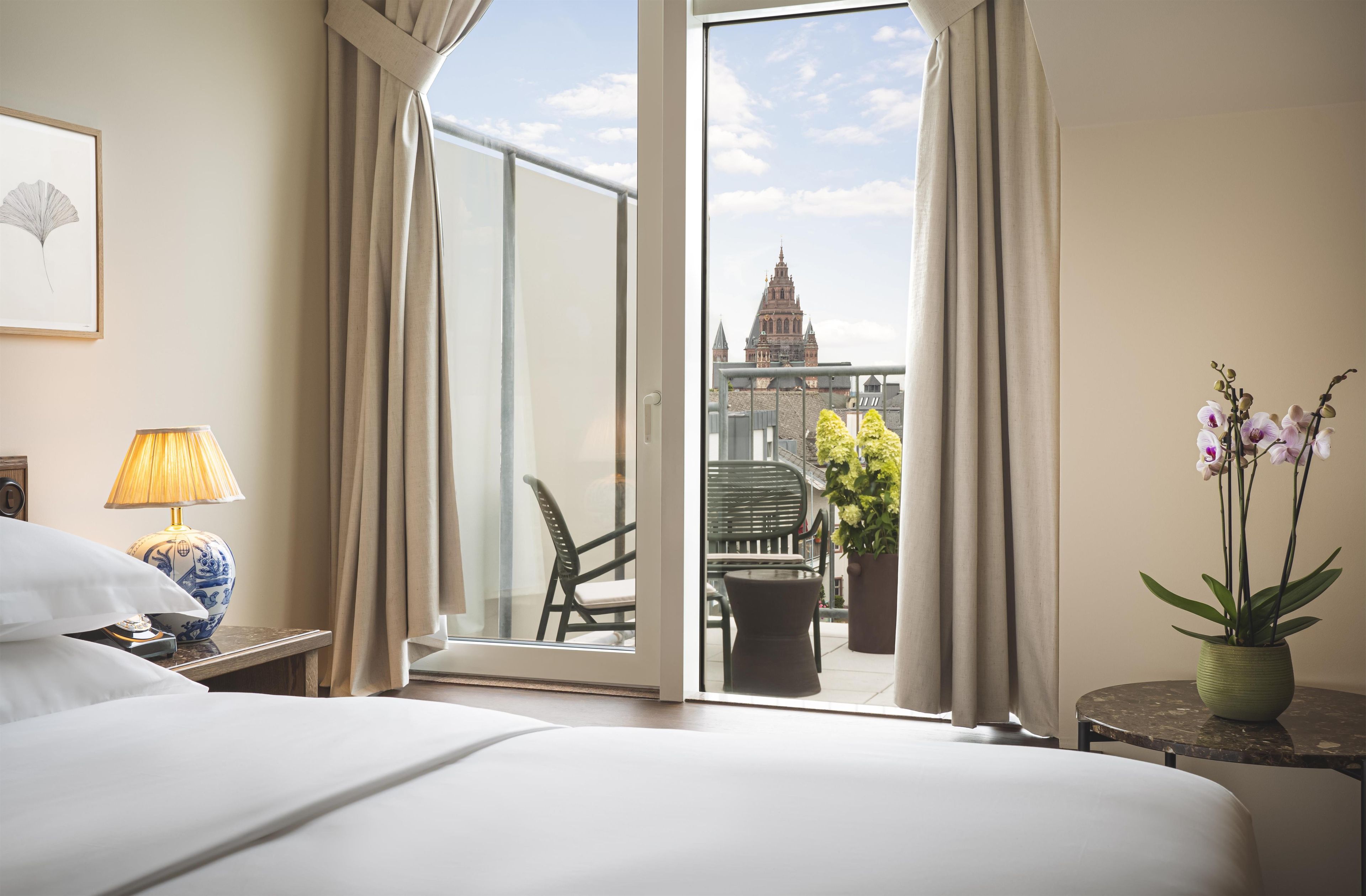 Una habitación de hotel con una cama, una lámpara, un florero con flores y vista a un edificio desde una puerta abierta.