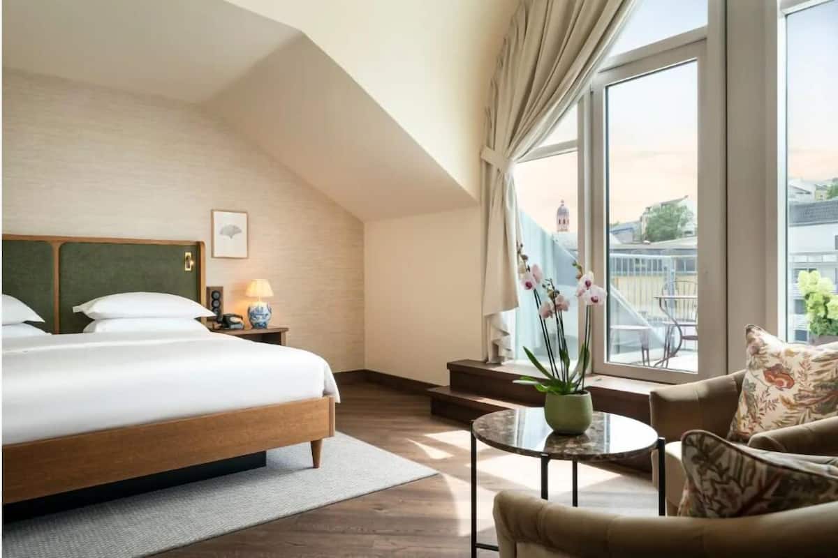 Habitación de hotel con una cama grande, una mesita con lámpara, un sofá y vistas a la ciudad a través de amplios ventanales.