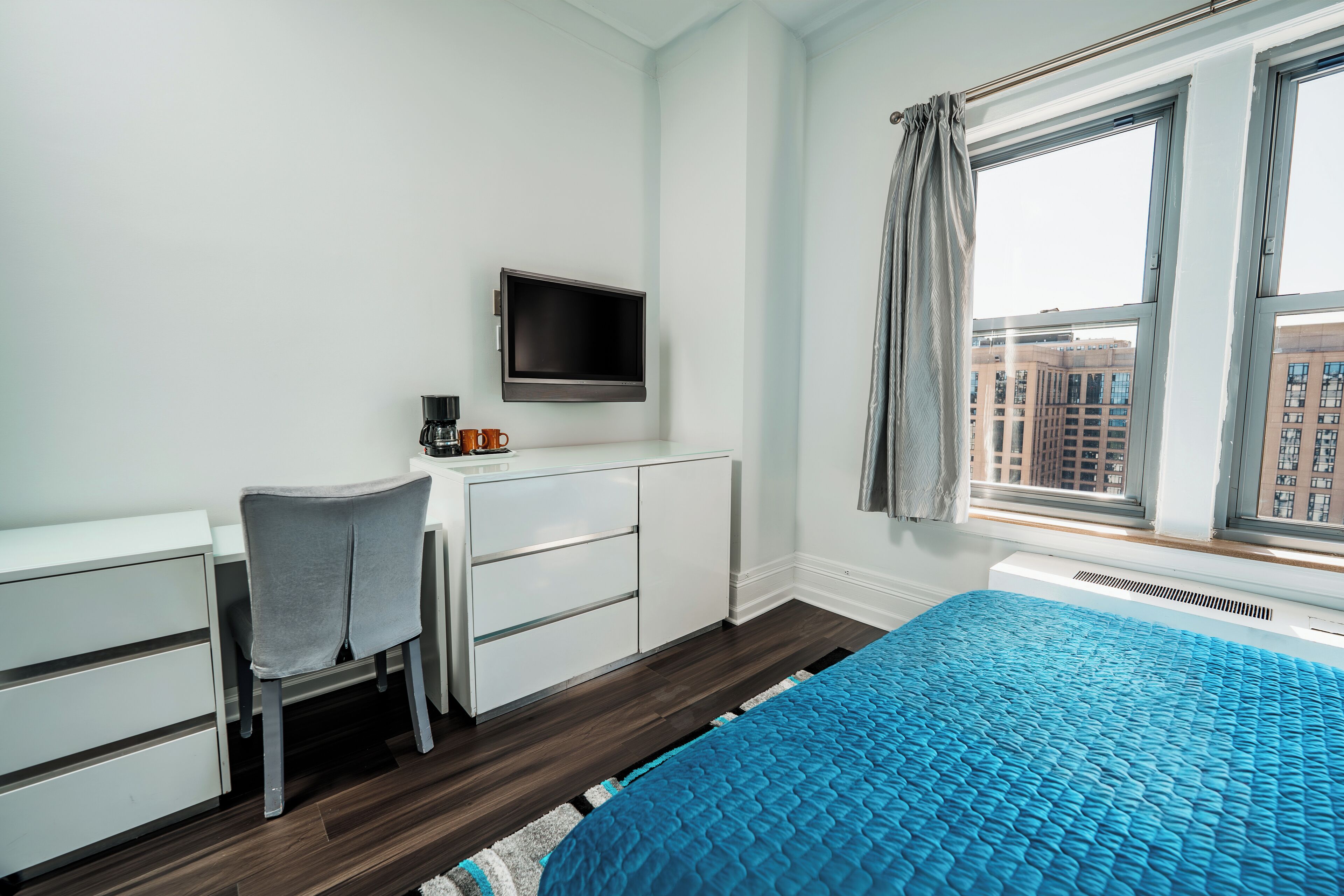 A bedroom with a bed, a desk with a chair, a television, and a window with a view of buildings.