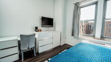 A bedroom with a bed, a desk with a chair, a television, and a window with a view of buildings.
