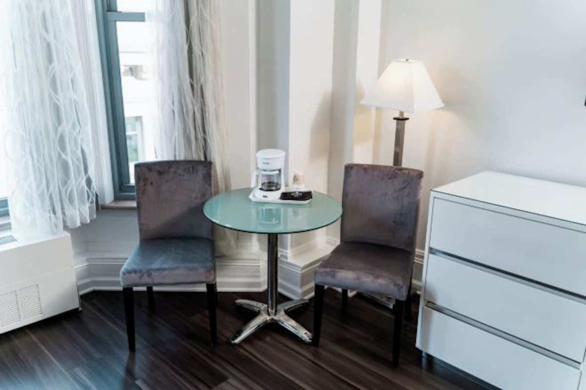 A hotel room with a glass table, two chairs, a lamp, a white cabinet, and a window with curtains.