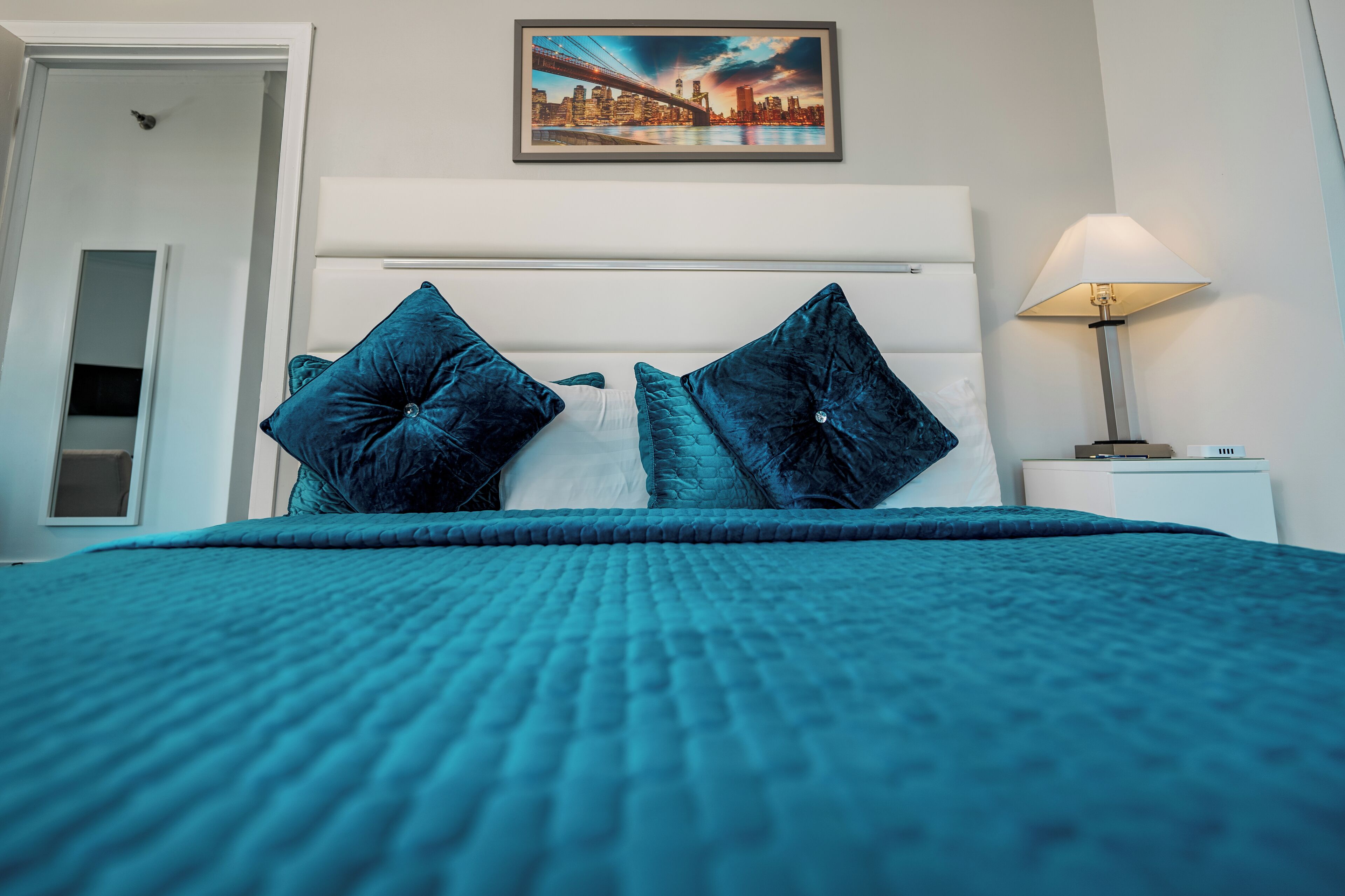 Una cama con una colcha color turquesa y dos almohadas azul oscuro, un cuadro enmarcado con una vista urbana en la pared, y una lámpara de noche.