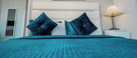 A bed with a teal bedspread and two dark blue pillows, a framed cityscape picture on the wall, and a bedside lamp.